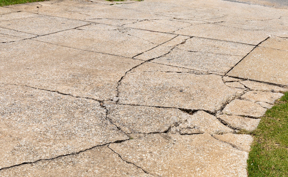 Cracked and broken aggregate concrete driveway descending into an asphalt roadway